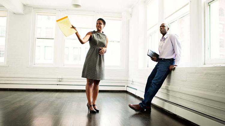 Man and woman standing in an empty apartment with dark hardwood floors and large windows along two walls..